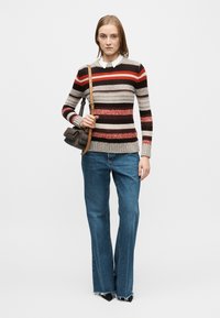 Prugasti pleteni džemper u sivoj, crvenoj i smeđoj boji. Kombiniran s lagano plavim trapericama širokih nogavica. Model drži malu tamnu torbu s naramenicom.