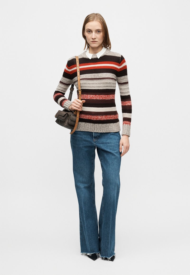 Prugasti pleteni džemper u sivoj, crvenoj i smeđoj boji. Kombiniran s lagano plavim trapericama širokih nogavica. Model drži malu tamnu torbu s naramenicom.