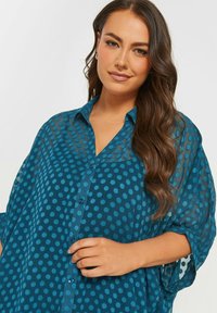 Camicia trasparente color turchese con grande motivo a pois circolari, chiusura a bottoni sul davanti e maniche svasate, caratterizzata da una texture leggermente traslucida.