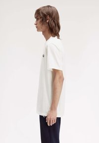 Weißes Baumwoll-T-Shirt mit kurzen Ärmeln und Rundhalsausschnitt, versehen mit einem kleinen schwarzen Logo auf der linken Brust. Glatte Textur, lässige Passform.