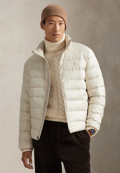 Doudoune de couleur crème clair, modèle zippé, poignets côtelés, portée sur un gros pull en maille torsadée beige, un pantalon en velours côtelé marron et un bonnet marron clair.