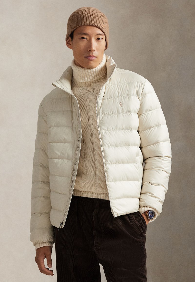 Šviesios kreminės spalvos pūkinė striukė su užtrauktuku, sukirpimas su ribinėtomis rankogaliais, dėvima ant didelio storos megztos smėlio spalvos megztinio, rudų kordurojų kelnių ir rudos spalvos vilnonės kepurės.