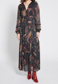 Pitkä musta kukallinen mekko paisley-kuvioilla, kerätty vyötärö ja läpinäkyvät pitkät hihat. Pukeutunut burgundinpunaisiin korkokenkiin.