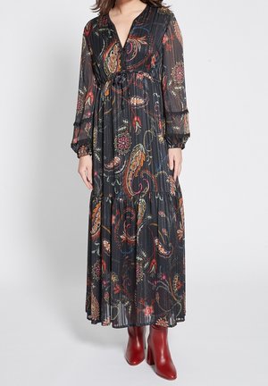 Lange zwarte bloemenjurk met paisleypatronen, verstelbare taille en doorschijnende lange mouwen. Gekleed met bordeauxrode haklaarzen.