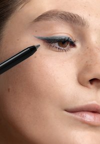 Un crayon eyeliner noir est appliqué près de la paupière supérieure. L'œil présente un eyeliner gris avec une aile. La peau a une texture lisse et uniforme avec des taches de rousseur.