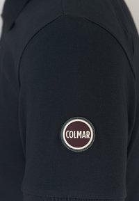 Polo navy in tessuto testurizzato, con un patch circolare del logo Colmar in bordeaux e argento sulla manica sinistra.