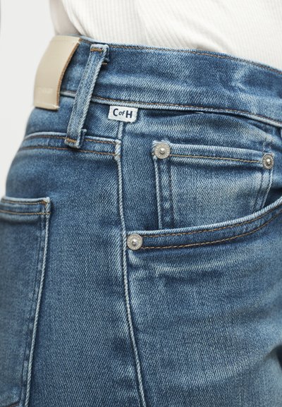 Κοντινό πλάνο στην πίσω τσέπη ενός μπλε τζιν με μεταλλικά καρφιά και μικρή λευκή υφασμάτινη ετικέτα με την ένδειξη «C of H.»