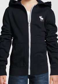 Zwarte zip-up hoodie van zachte stof, met een witte rits en een klein wit logo van een eland op de linkerborst. Met capuchon.