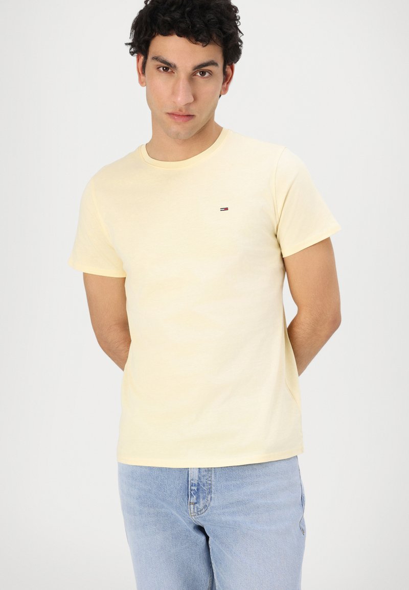 Camiseta de algodón amarilla clara con cuello redondo, mangas cortas y un pequeño logotipo en el pecho. Combinada con jeans azul claro.