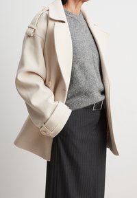 Personne portant un manteau beige oversize, un pull gris texturé et une jupe noire à fines rayures avec une ceinture noire, debout devant un fond uni.