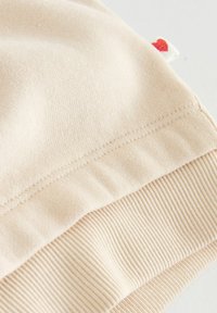 Beiger Sweatshirtstoff aus Baumwolle mit glatter Oberfläche und geripptem Saum. Ein auffälliges Detail ist ein kleines rotes Herzetikett an der Seite.