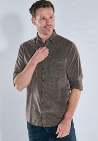 Braunes Hemd mit Knöpfen aus strukturiertem Stoff, hochrollbaren Ärmeln, einer Brusttasche und dezenten Nähdetails. Getragen mit dunkelblauen Jeans.