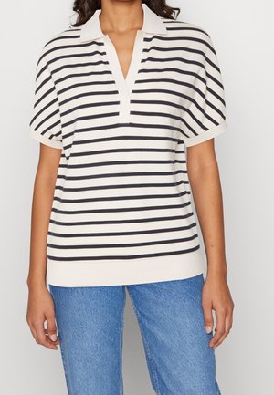 Nainen, joka on pukeutunut mustavalkoraidalliseen lyhythihaiseen poolopaitaan ja sinisiin farkkuihin, yksiväristä taustaa vasten.