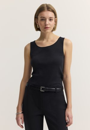 Frau mit kurzen blonden Haaren, die ein ärmelloses schwarzes Oberteil, schwarze Hose, schwarzen Gürtel, goldene Creolen und ein silbernes Armband trägt und nach vorne schaut.