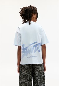 Lys blå kortærmet T-shirt med et stort graffiti-lignende print på ryggen med teksten "SANT." og "SET PÅ GADERNE."