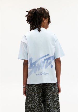 Hellblaues T-Shirt mit kurzen Ärmeln, das auf dem Rücken mit einem großen Graffiti-ähnlichen Druck versehen ist, auf dem die Texte "SANT." und "AS SEEN ON THE STREETS." zu sehen sind.