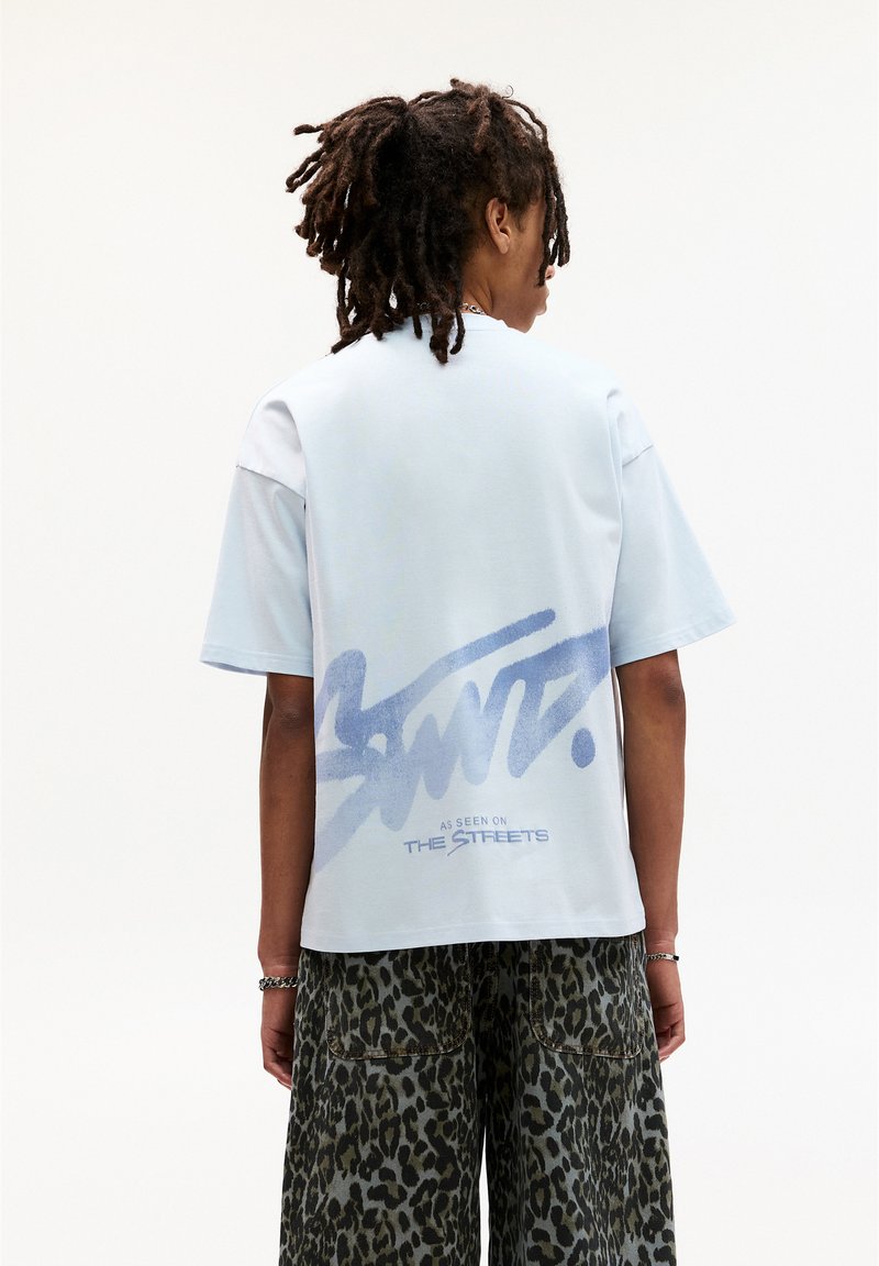 Lys blå kortærmet T-shirt med et stort graffiti-lignende print på ryggen med teksten "SANT." og "SET PÅ GADERNE."