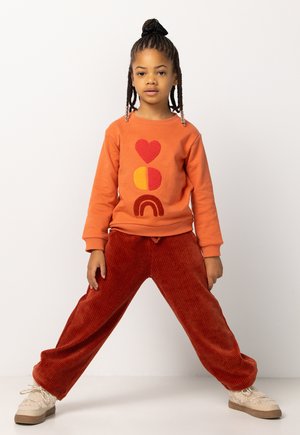 Oranger Sweatshirt mit Herz-, Kreis- und Regenbogenmotiven aus plüschigem Garn, kombiniert mit weit geschnittenen rostfarbenen Cordhosen und beigen Schuhen.
