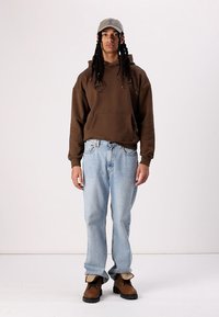 Sweatshirt com capuz castanho, jeans azuis e botas castanhas. Boné cinza. Capuz com cordões, dois bolsos frontais, ajuste relaxado, jeans de lavagem clara.