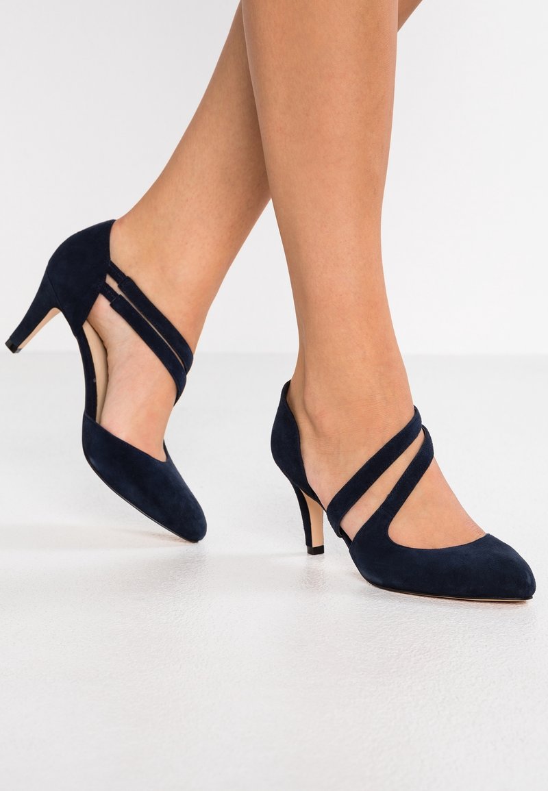 Pieds portant des talons hauts en daim bleu marine avec deux brides diagonales sur le dessus, debout sur un sol blanc.