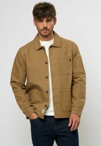 Chaqueta de lona marrón con cuello tipo solapa, cierre de botones y dos bolsillos frontales. Llevada sobre un suéter blanco texturizado.