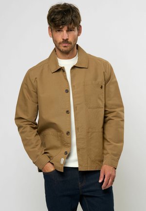 Veste en toile marron avec un col, fermeture à boutons et deux poches avant. Portée par-dessus un pull blanc texturé.