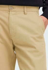 Chino en coton beige avec une texture lisse, comportant une poche latérale et un bouton décoratif avec une finition neutre.
