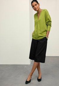 Grüner Wollcardigan mit V-Ausschnitt, Fronttaschen und Knopfverschluss, kombiniert mit schwarzen Culottes und grünen Wildlederabsätzen.