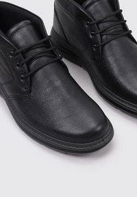 Un par de botas negras de cuero con cordones hasta el tobillo y punteras redondeadas sobre un fondo gris claro.