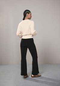 Woman with braided hair wearing a cream sweater, black flared pants, and metallic flat shoes standing against a plain grey background.