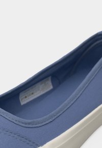 Gros plan sur une chaussure bleu toile sans lacets avec une semelle en caoutchouc blanche et une étiquette intérieure visible sur un fond uni.