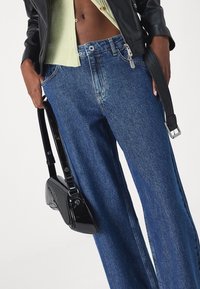 Široké modré džíny s vysokým pasem, vyrobené z denimu, doplněné lesklou černou kabelkou přes rameno s zakřiveným designem a nastavitelným popruhem.