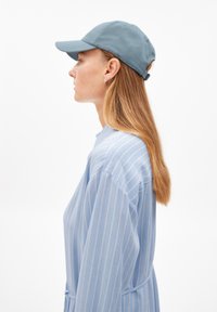 Chemise à rayures bleues à coupe ample, avec des manches longues et un nœud à la taille, associée à une casquette de baseball bleu clair.