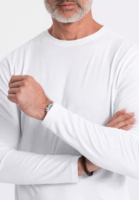 Pitkähihainen valkoinen paita, joka on valmistettu pehmeästä kankaasta ja jossa on pyöreä pääntie. Rannekellon näkyminen ranteessa, kädet ristissä rentoon ja mukautuvaan ilmeeseen.