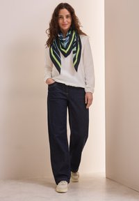 Blusa bianca a maniche lunghe, pantaloni ampi in denim scuro e una sciarpa di seta multicolore con motivi geometrici in verde, blu e nero.