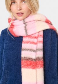 Vilnonis šalikas su rožinėmis, koralų ir pilkomis juostomis ant minkštos tekstūros, užmestas ant kaklo, derinamas su tamsiai mėlynu pūkuotu megztiniu.