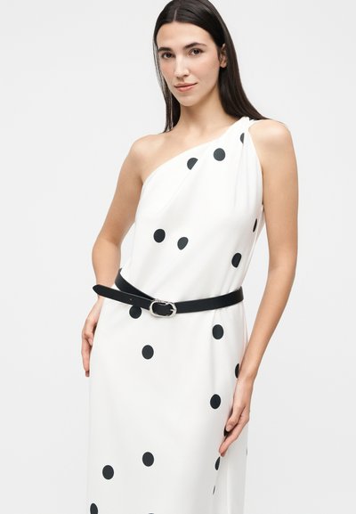 Γυναίκα που φοράει λευκό φόρεμα με έναν ώμο, μαύρες πουά και μαύρη ζώνη, στέκεται με το ένα χέρι στη μέση.