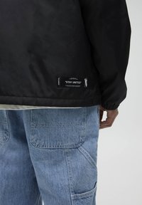 Czarna kurtka o gładkiej fakturze, z elastycznymi mankietami i metką "STAY UNITED". W komplecie jasnoniebieskie dżinsy z kieszeniami.