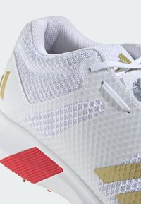 White athletic shoe with a textured mesh upper, gold accents, and red outsole detailing. Features traditional lacing and padded collar.
