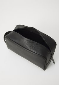 Trousse de toilette en cuir noir avec surface texturée, de forme rectangulaire et fermeture éclair supérieure. L'intérieur est spacieux et dépouillé.