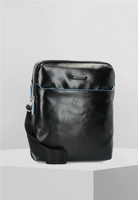 Czarna skórzana torba o gładkiej fakturze, z niebieskim akcentem w postaci zamka błyskawicznego, regulowanym paskiem oraz dyskretnym logo z przodu.