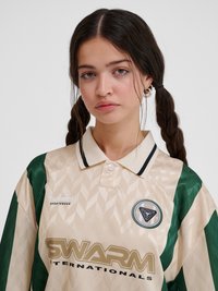 Noor naine, kellel on pikk tume punutud soeng, kannab beežikat ja rohelist spordisärki logode ja kraega, seisab ühevärvilise tausta ees.