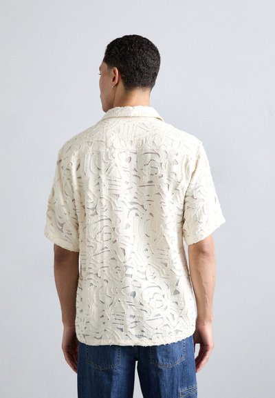 Kortsleeve skjorte i kremfarget stoff med teksturerte utskjæringsmønstre, en avslappet passform og en camp-stil krage, båret med blå denimjeans.