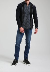 Giacca a zip blu sopra una camicia con motivo, abbinata a jeans blue e sneakers nere con suole bianche. Materiali texturizzati visibili.