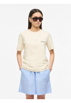 Mujer con cabello largo y oscuro que lleva gafas de sol negras extragrandes, camiseta crema con logo y pantalones cortos azul claro con rayas finas, con las manos en los bolsillos.