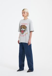 Šedé oversized tričko s barevným potiskem tygra a textem, kombinované s volnými modrými džínami a černými špičatými botami s akcentními detaily.