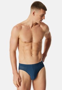 Marineblaue Badebriefs mit schmalen vertikalen Streifen, ausgestattet mit einer glatten Textur und einem elastischen Bund. Minimale Markenkennzeichnung an der Seite.