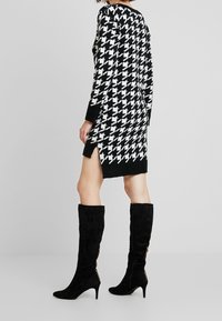 Černobílé pletené šaty s kostkovaným vzorem houndstooth a postranními rozparky, doplněné černými zamšovými kozačkami po kolena s akcenty zlatého zipu.
