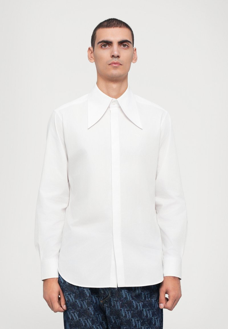 Camisa blanca de manga larga con un gran cuello puntiagudo, que presenta una tapeta de botones en el frente y un sutil logo bordado en el pecho.