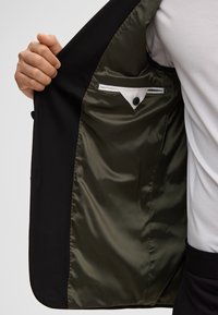 Mano che tiene aperta una giacca nera con fodera verde oliva lucida, mostrando la tasca interna con patta triangolare bianca e bottone nero.